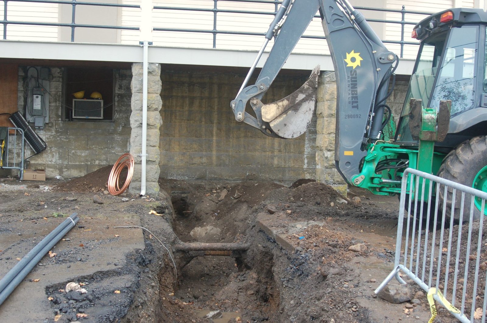 A backhoe bucket over a muddy trench beside a building, with exposed pipe