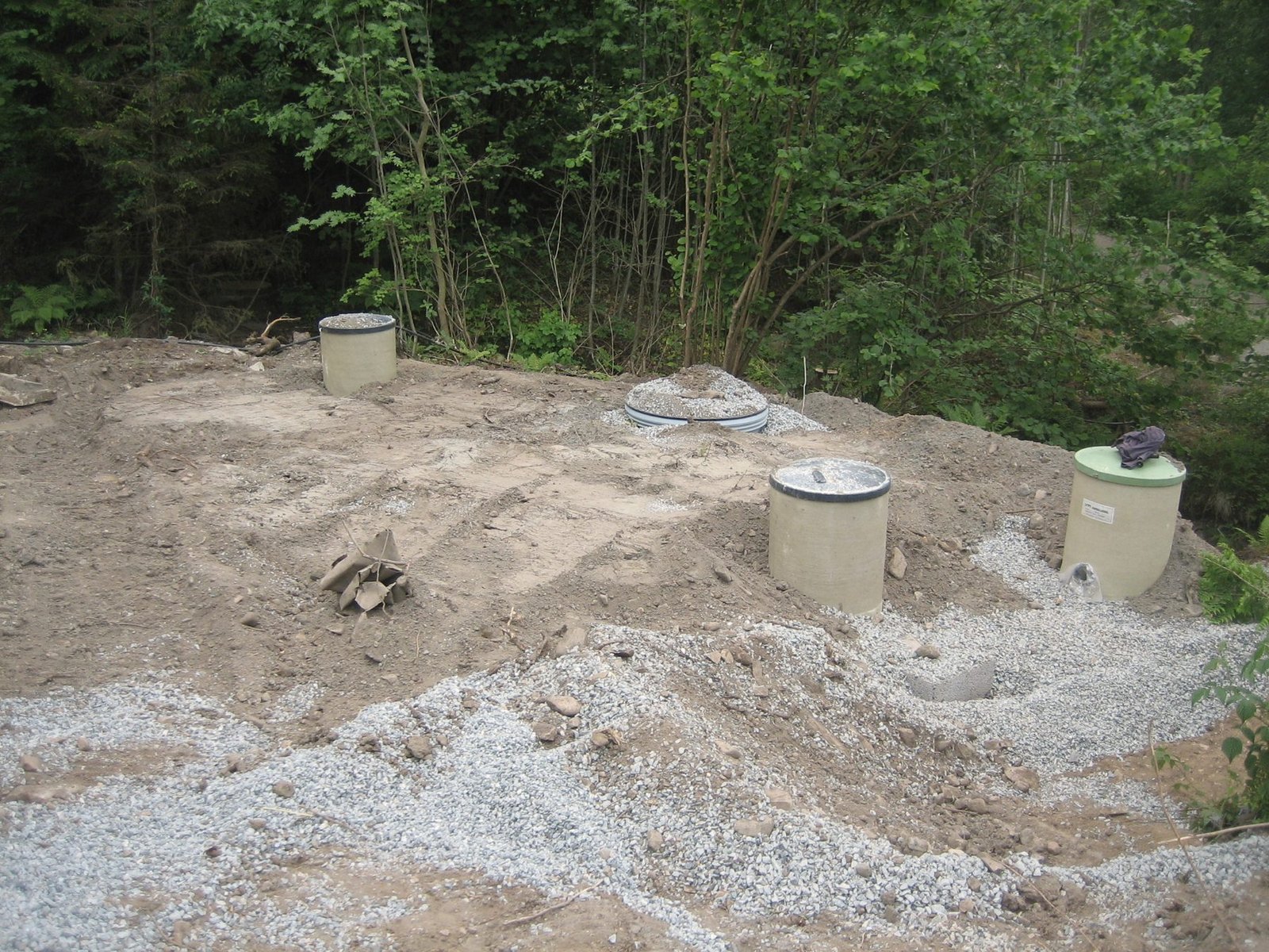Three septic tank risers standing in freshly graded sand and gravel at the edge of woods